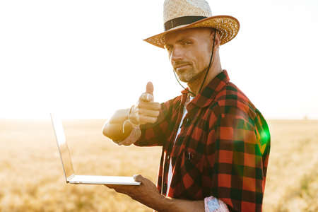 Image of unshaven adult man in straw hat working with laptop while pointing finger at camera at cereal fieldの写真素材