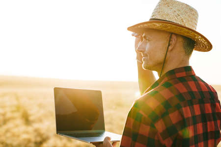 Image of pleased adult man in straw hat working with laptop while standing at cereal fieldの写真素材