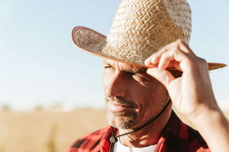Image of unshaven adult man in straw hat looking aside while standing at cereal fieldの写真素材