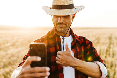 Image of pleased adult man in straw hat taking selfie on cellphone while showing thumb up at cereal fieldの写真素材