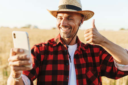 Image of smiling adult man in earphones showing thumb up and taking selfie on cellphone at cereal fieldの写真素材