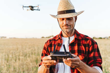 Image of pleased handsome man in straw hat using drone while standing at cereal field