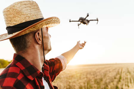 Image of focused handsome man in straw hat using drone while standing at cereal fieldの写真素材