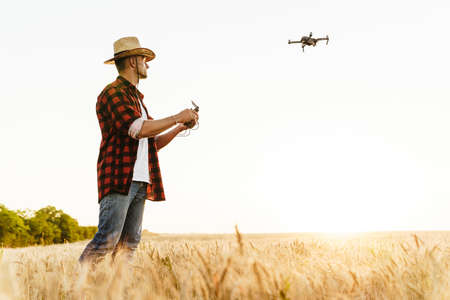 Image of focused handsome man in straw hat using drone while standing at cereal fieldの写真素材