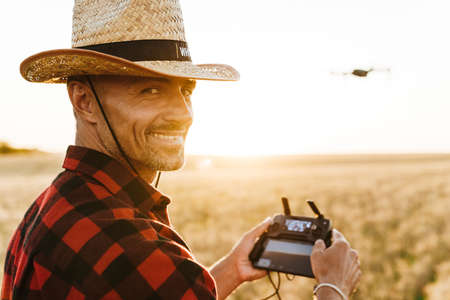 Image of happy handsome man in straw hat using drone while standing at cereal fieldの写真素材