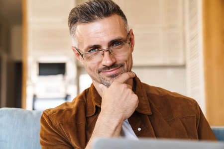 Image of smiling adult man in eyeglasses working with laptop while sitting on couch at homeの写真素材