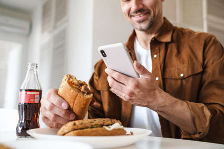 Cropped image of joyful man smiling and using smartphone while have lunch in white kitchenの写真素材