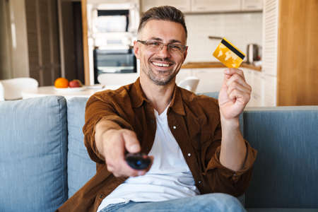 Image of handsome smiling man holding credit card and using remote control while watching tv at home