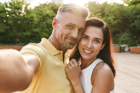 Portrait of happy middle-aged couple man and woman taking selfie photo and hugging while walking in...