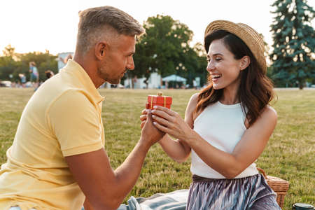 Image of happy adult man giving present box to beautiful woman while sitting on grass in summer park