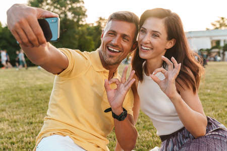 Portrait of smiling adult couple man and woman gesturing ok sign and taking selfie photo on...