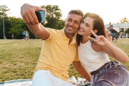Portrait of smiling adult couple man and woman gesturing peace sign and taking selfie photo on...