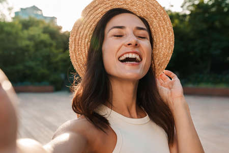 Image of nice middle-aged woman wearing straw hat taking selfie photo and laughing while walking in summer parkの写真素材