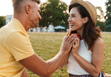 Image of smiling adult man giving present box to beautiful woman while sitting on grass in summer...