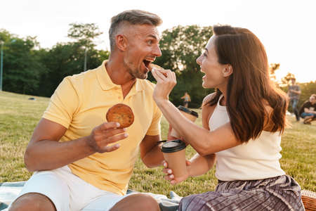 Portrait of delighted middle-aged couple man and woman drinking coffee takeaway and eating cookies while sitting on grass in parkの写真素材