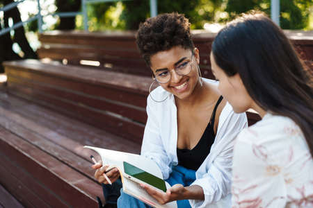Image of cheerful african positive woman with her friend doing homework outdoors in park while using...