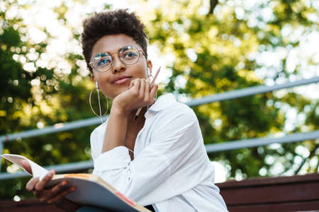 Photo of positive concentrated african woman sitting outdoors in park while listening music and...