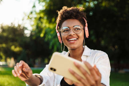 Photo of happy smiling african woman sitting outdoors in park on a grass while listening music with headphones and using mobile phoneの写真素材