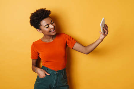 Image of happy african american woman taking selfie photo on mobile phone isolated yellow background