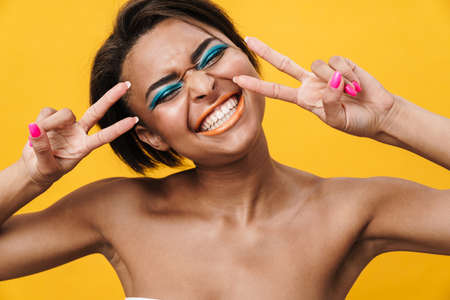 Image of shirtless african american woman laughing while gesturing peace sign isolated over yellow...