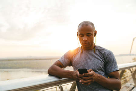 Sporty young african man typing on a smart phone while resting outdoors, on the bridgeの写真素材