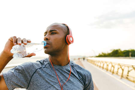 Young african sports man relaxing after jogging, holding water bottle on a bridge, wearing headphonesの写真素材
