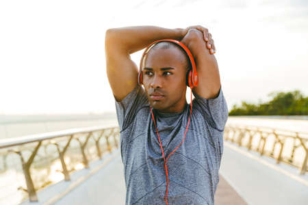 Relaxed african sportsman stretching while standing on the bridge, wearing headphonesの写真素材