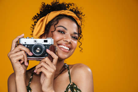 Image of cheerful african american woman taking photo on retro camera isolated over yellow wall