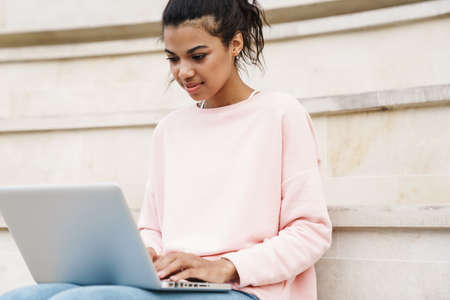 Image of focused african american student girl working with laptop while sitting on stairs outdoorsの写真素材