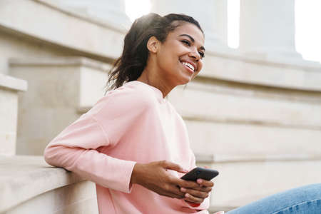 Image of smiling african american girl using cellphone while sitting on stairs outdoors