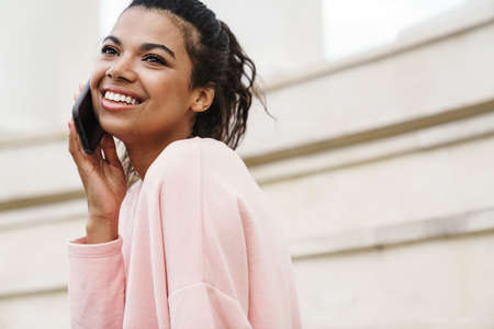 Image of smiling african american girl using cellphone while sitting on stairs outdoors