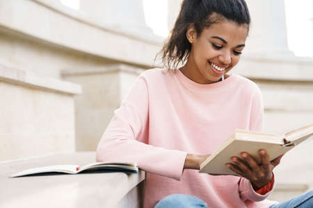 Image of cheerful african american student girl reading book while sitting on stairs outdoorsの写真素材