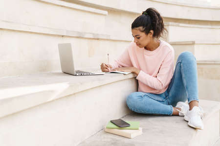 Image of pleased african american student girl doing homework with laptop while sitting on stairs...