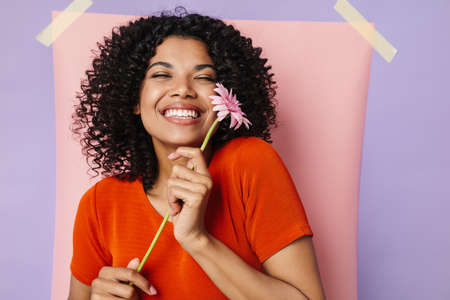Image of cheerful african american woman smiling while posing with flower isolated over multicolored...