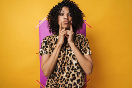 Image of amusing african american woman pointing fingers at cheeks isolated over multicolored...