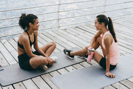 Image of joyful multinational sportswomen talking and drinking water while sitting on mats at promenadeの写真素材