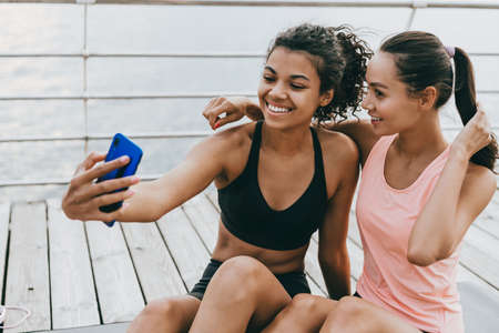 Image of happy multinational sportswomen taking selfie on cellphone while sitting on promenadeの写真素材