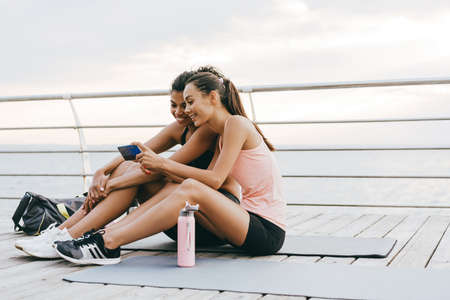 Image of smiling multinational sportswomen using cellphone while sitting on promenade