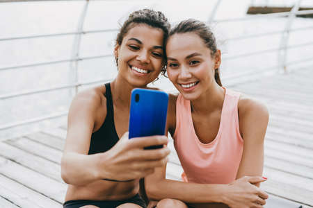 Image of cheerful multinational sportswomen taking selfie on cellphone while working out at promenadeの写真素材