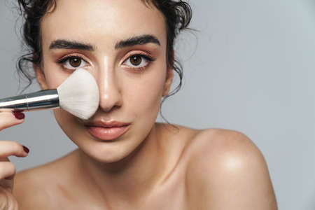 Image of beautiful half-naked woman using powder brush on camera isolated over gray wall