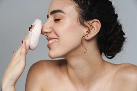 Image of pleased half-naked woman smiling and using powder sponge isolated over gray wallの写真素材