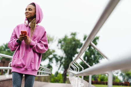 Attractive young african sports woman listening to music with headphones in the park, holding mobile phoneの写真素材
