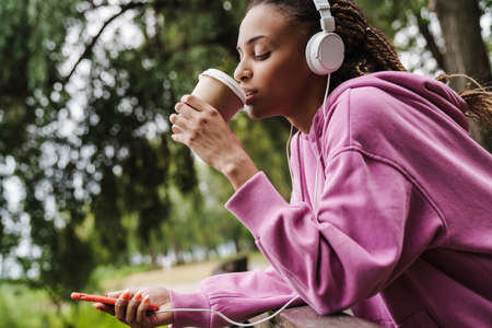 Attractive young african sports woman listening to music with headphones in the park, holding mobile phone, drinking takeaway coffeeの写真素材