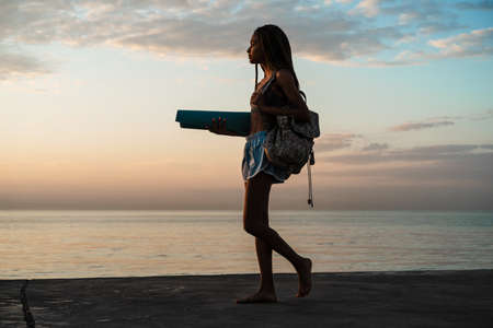 Image of calm african american girl in sportswear walking with mat on concrete promenade outdoors