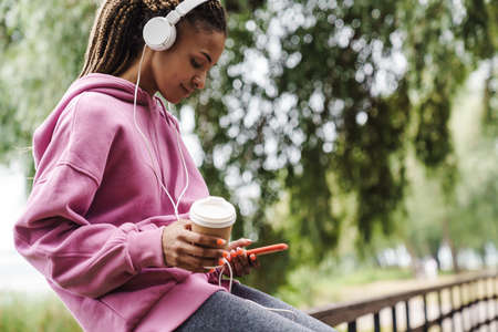 Attractive young african sports woman listening to music with headphones in the park, holding mobile phone, drinking takeaway coffeeの写真素材