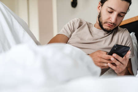 Tired sleepy young man using mobile phone while laying in bed in the morningの写真素材