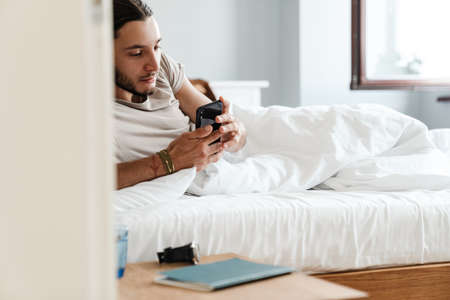 Smiling young man using mobile phone while laying in bed in the morningの写真素材