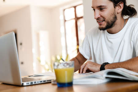 Confident young man working on laptop computer while sitting at the table at home, writing notes