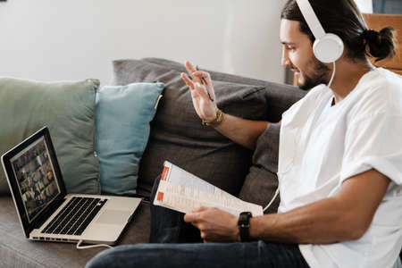 Confident young man wearing headphones having a video meeting via laptop while sitting on a couch at...