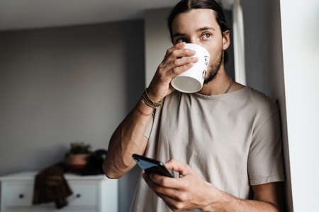 Attractive young guy using mobile phone standing at home, holding cupの写真素材